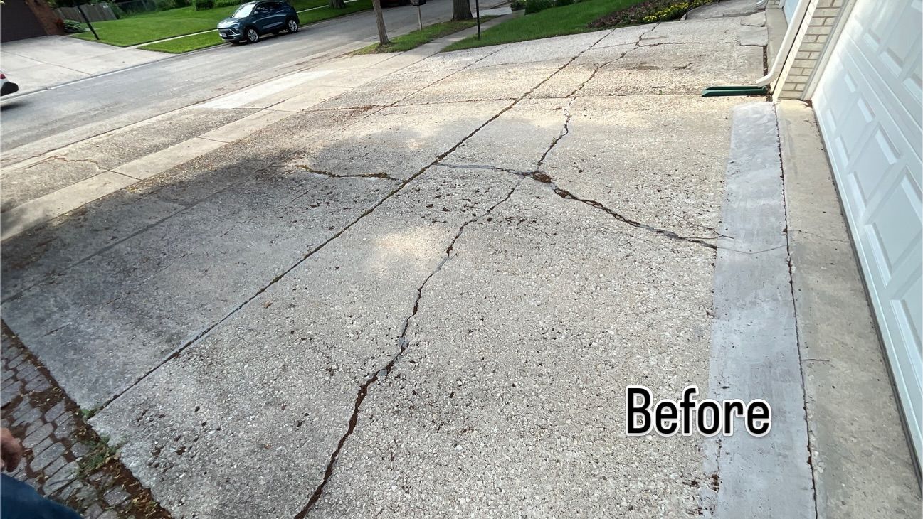 An old driveway with long cracks, pitting and a patched strip by the garage